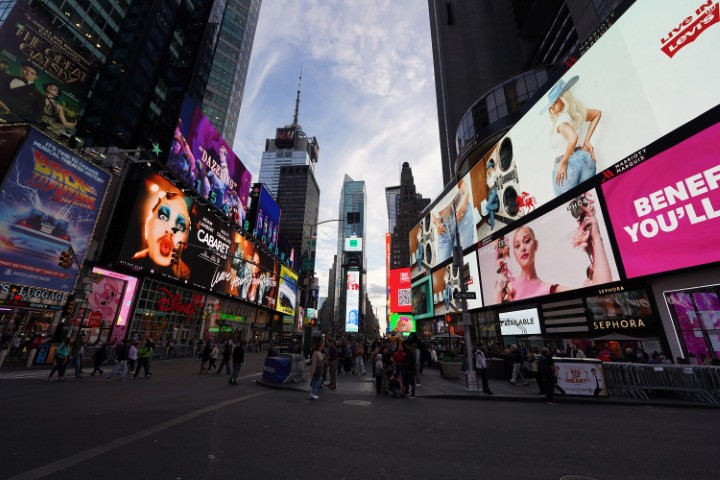 One of the streets in Times Square. Many people are walking, and buildings with huge advert screens are on either sides of the street.